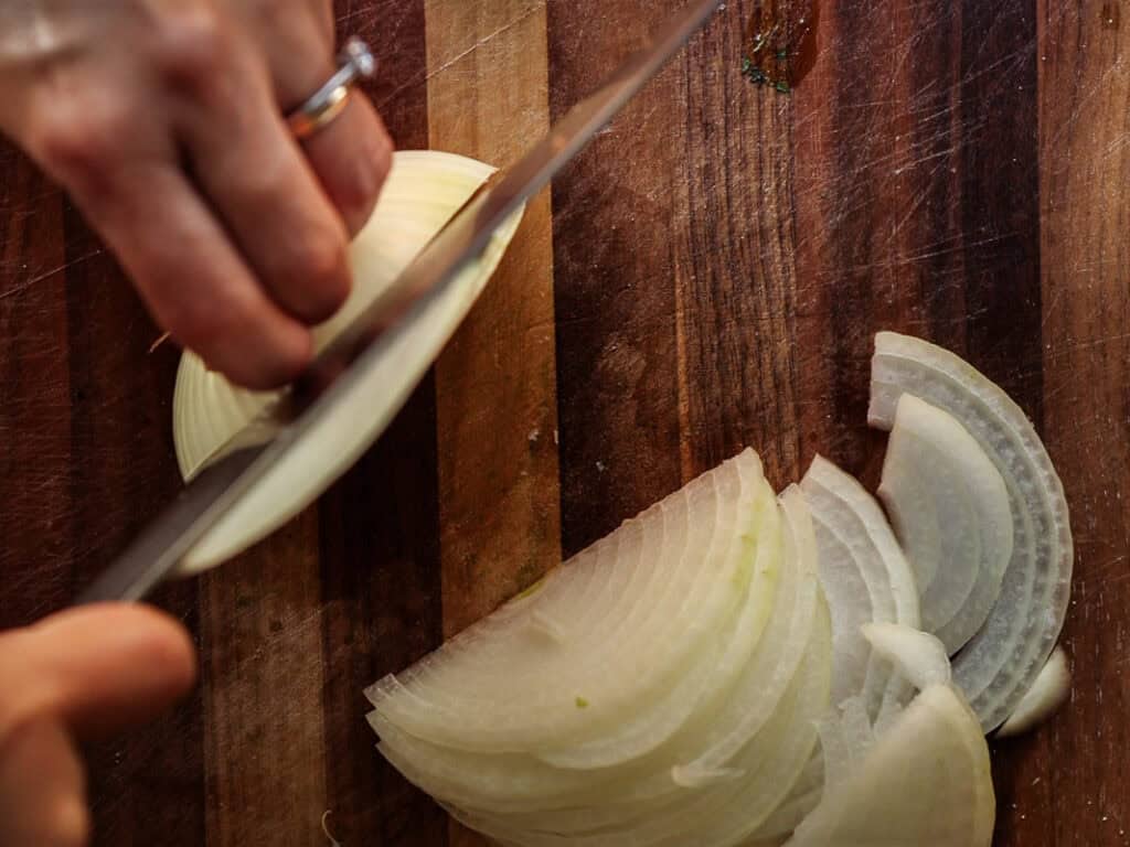 A person slices an onion on a wooden cutting board. The left hand holds the onion steady, while the right hand uses a knife to cut even slices. The onion pieces are arranged on the cutting board.